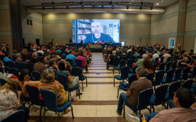 Foto do plenário do evento, tendo ao fundo a tela onde aparece o jornalista Jamil Chade