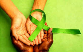 Imagem composta de um laço verde repousando sobre as palmas das mãos de uma pessoa branca. essas mãos estão sobre as palmas das mãos de uma pessoa negra. o fundo da imagem é verde