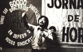 Imagem mostra Luiz Gushike, ex-presidente do Sindicato dos Bancários de São Paulo, Osasco e Região. ele é um homem oriental e, na foto, está na casa dos 30 anos. Veste camisa e gravata e fala ao microfone, com a mão esquerda levantada