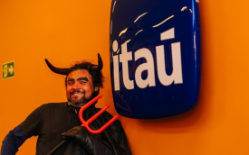 Imagem mostra homem fantasiado de diabo apontando um tridente para o logo do Itaú pendurado em uma parede pintada de laranja