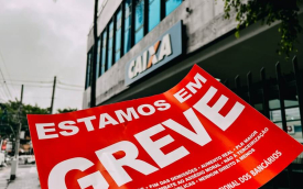 Imagem mostra um adesivo na cor vermelha onde se lê "Estamos em Greve" em letras brancas e, ao fundo, uma agência da Caixa Econômica Federal