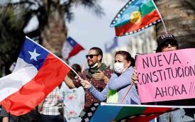 Foto mostra manifestantes chilenos, durante protesto. Um deles carrega uma bandeira chilena, outra segura a bandeira mapuche, e uma terceira pessoa segura um cartaz onde se lê, em espanhol: "nova constituição agora"
