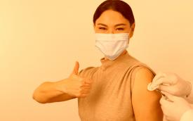 Imagem mostra uma mulher de origem asiática sendo vacinada. Ela faz um sinal de positivo com a mão esquerda