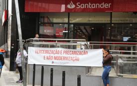 Imagem de protesto em frente a fachada de agência do Santander. Dois manifestantes seguram uma faixa onde se lê 'terceirização é precarização e não modernização'