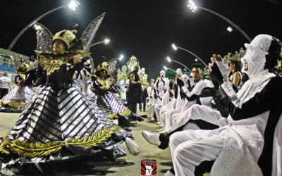 Fotografia mostra integrantes fantasiados de preto e branco ajoelhados ao lado de uma porta-bandeira da Vai-Vai
