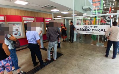 Imagem mostra área de autoatendimento de agência do Bradesco durante protesto do Sindicato contra demissões e assédio moral. Na imagem, diversos clientes e os dirigentes portando uma faixa onde se lê "pelo fim do assédio moral"