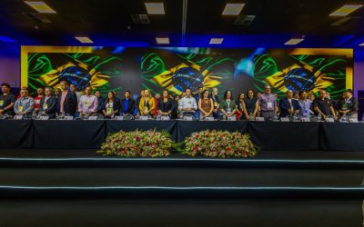 Imagem mostra palanque da abertura da 27ª conferência nacional dos bancários. vários dirigentes sindicais bancários estão perfilados lado a lado diante de uma mesa comprida. Um telão exibe três bandeira do Brasil, lado a lado