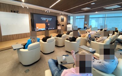 Imagem mostra uma sala cheia de pessoas. Elas estão sentadas em poltronas e de frente para um pequeno palco