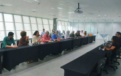 Imagem mostra sala onde ocorreu a reunião para discutir o Saúde Caixa. a imagem mostra 12 pessoas sentadas lado a lado em uma mesa comprida. Atrás dela há uma janela que ocupa toda a parede.