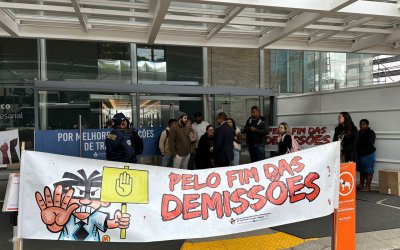 Imagem mostra protesto em frente a uma agência do Itaú, com uma faixa exposta em primeiro plano onde se lê "pelo fim das demissões"