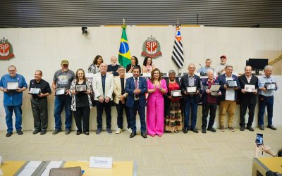 Imagem mostra personagens que fizeram a greve de 1985. Eles estão lado a lado na sala da assembleia legislativa de são paulo e seguram placas de homenagem