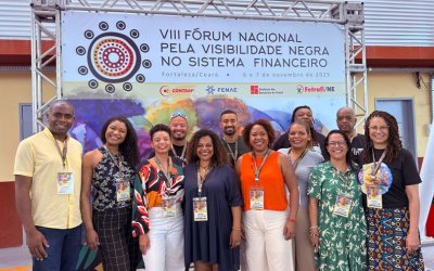 Imagem mostra membros do coletivo de combate ao racismo do Sindicato dos Bancáiros de São Paulo em frente a um painel do VIII Fórum Nacional pela Visibilidade Negra no Sistema Financeiro