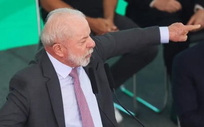 Imagem mostra o presidente da República, Luiz Inácio Lula da Silva. Ele um homem de pele, cabelos e barba branca. Veste um terno preto, camisa azul clara e gravata rosa. Seu braço direito está erguido na altura do ombro