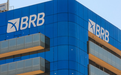 Imagem mostra o alto da fachada do prédio do BRB. o edifício é espelhado com vidros azuis. a imagem foca no lato da fachada, que exibe o logo do banco