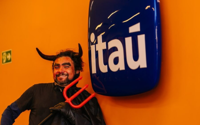 Imagem mostra homem fantasiado de diabo apontando um tridente para o logo do Itaú pendurado em uma parede pintada de laranja