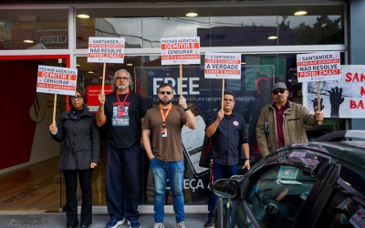 imagem mostra dirigentes perfiladas portando placas em frente a uma agência do Santander durante protesto
