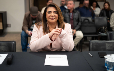 Neiva Ribeiro, presidenta do Sindicato dos Bancários, veste um blazer branco e usa óculos. Está sentada, com os braços apoiados em uma mesa, e dedos das mãos entrelaçados, na altura do peito