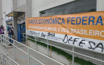 Fachada da agência da Caixa Federal no Parque São Domingos com um faixa do Sindicato estendida, escrita: Caixa Econômoca Federal, um patrimônio do Brasil e dos brasileiros. Na sua parte inferior, a faixa está pixada com a frase: Lutamos em sua defesa. Mais ao fundo da foto, em frente a sua porta, o dirigente André Sardão.