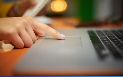 Foto mostra mão feminina mexendo em um laptop sobre uma mesa de madeira