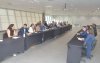 Imagem mostra sala onde ocorreu a reunião para discutir o Saúde Caixa. a imagem mostra 12 pessoas sentadas lado a lado em uma mesa comprida. Atrás dela há uma janela que ocupa toda a parede. 