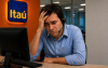 Imagem mostra bancário com semblante preocupado apoiando a cabeça na mão esquerda. Ele usa um headphone e uma camisa azul e está sentado em frente a um computador.  atrás dele há uma placa com o logo do Itaú pendurado em uma janela pintada de laranja