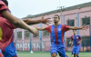Jogador de futebol após marcar um gol correndo para receber o abraço dos companheiros.