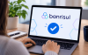 Imagem mostra uma mulher sentada em frente a um computador portátil em cuja tela é projetado um sinal de "check"  abaixo do logo do Banrisul