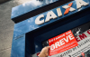 Imagem mostra uma mão segurando um adesivo onde se lê "greve", em frente à fachada de uma agência da Caixa