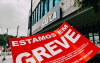Imagem mostra um adesivo na cor vermelha onde se lê "Estamos em Greve" em letras brancas e, ao fundo, uma agência da Caixa Econômica Federal