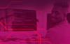 Imagem composta pela foto de um homem de pele negra sentado em frente a um computador, sob um filtro na cor roxa