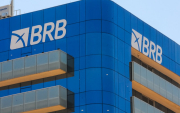 Imagem mostra o alto da fachada do prédio do BRB. o edifício é espelhado com vidros azuis. a imagem foca no lato da fachada, que exibe o logo do banco